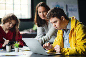 Student with down syndrome working at a laptop on HQIM. The teacher is looking over his shoulder and providing instruction based on training she received through CBPL