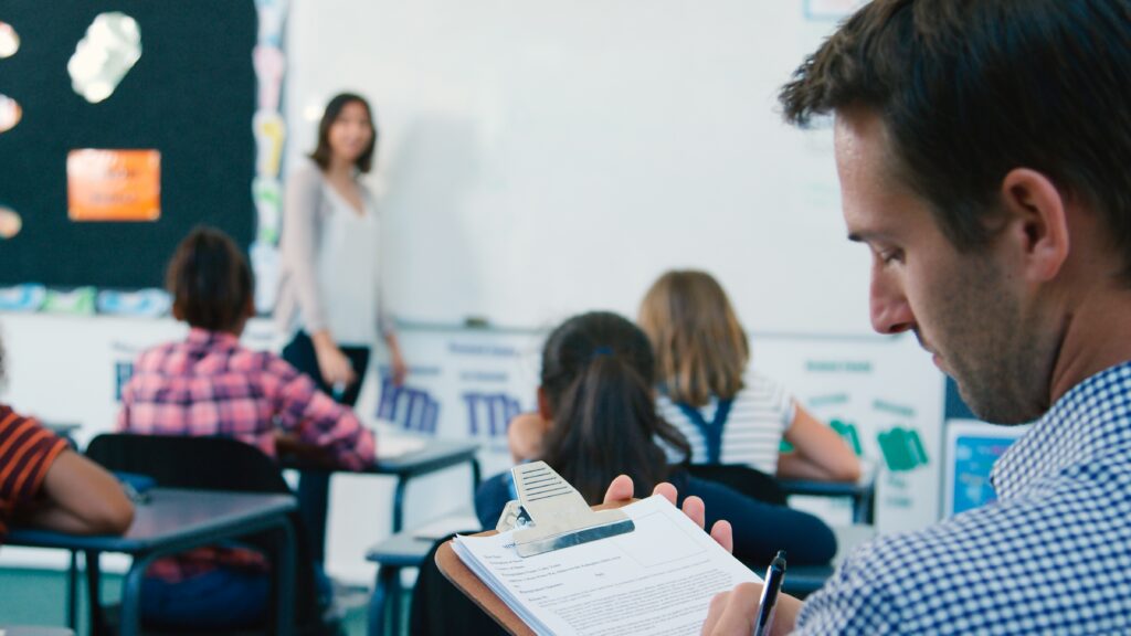 White male school leader observing a teacher. Writing observations about instructions on a clipboard.